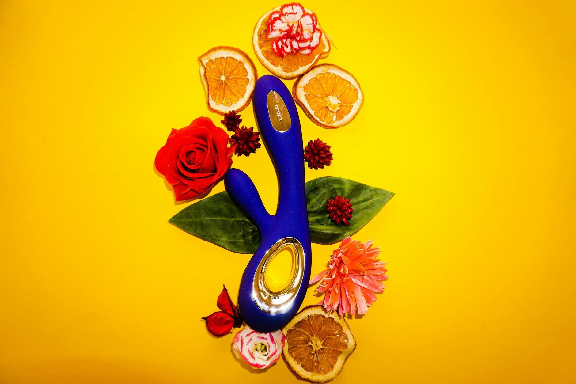 A blue vibrator surrounded by flowers and citrus slices on a yellow background, symbolizing healing and joy